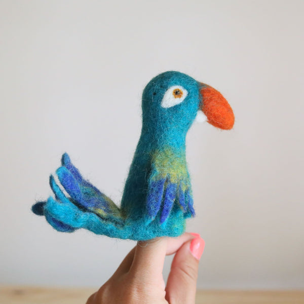 A handcrafted, three-dimensional parrot finger puppet made of wool felt is perched on one leg, displaying its vibrant blue body with green head and orange beak. The colorful puppet appears designed for children's play and storytelling, with a cheerful and crafty overall mood.