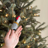A handmade felt penguin finger puppet with headphones is displayed by a female hand, set against a festive Christmas tree adorned with warm lights. The gray and white penguin features black accents, bright red headphones, and a yellow band, conveying a hip and playful attitude perfect for kids.