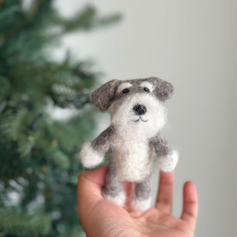 A handcrafted gray and white Schnauzer dog felt finger puppet is held gently in a female hand, set against a softly blurred Christmas tree background. This textured wool felt Schnauzer finger puppet features the breed's signature bushy eyebrows and beard, making it perfect for imaginative play.