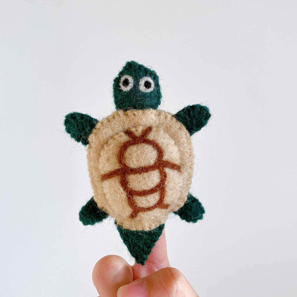 A hand holds a small, handmade turtle finger puppet resting on a plain white background. The felt turtle has a brown shell with lighter brown patterns resembling real turtle texture, and its head and limbs are in shades of brown with a simple smile design. The wool felt material is visible, and the 3D design makes it appealing from any angle.