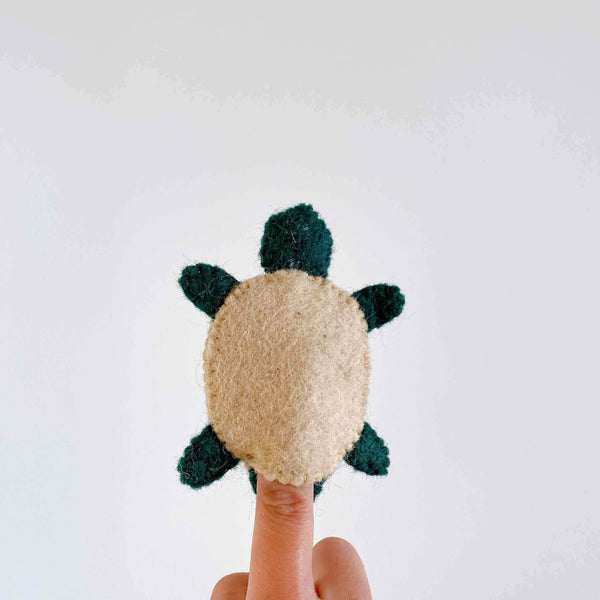 A close-up of a small, green turtle felt finger puppet resting against a plain background. The turtle has a brown felt bottom, textured shell, and smiling face. It's made from 100% wool felt, with a soft and plush appearance.
