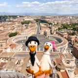 Two handmade felt duck finger puppets, depicting a duck couple on honeymoon, are held by a hand against a bright cityscape. These charming handcrafted felt ducks, made in Nepal, feature intricate wedding attire. The groom duck puppet wears a black hat and tie, while the bride duck puppet has a pink floral headpiece and a white pearl necklace. Both white felt ducks have orange beaks and feet.