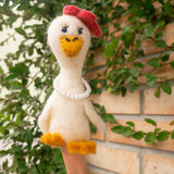 A handcrafted needle felted duck finger puppet, part of a 'Duck Couple on Honeymoon' set, is presented by a female hand against a backdrop of green foliage and a brick wall. This charming cream-bodied duck features a yellow beak and feet, a red bow, and a delicate pearl necklace, exuding a whimsical charm for imaginative storytelling.
