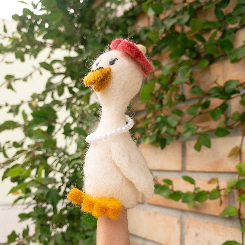 A handcrafted wool felt duck finger puppet is held by a female hand, set against lush green vines climbing a brick wall. This soft, off-white duck features a bright orange beak and feet. A red beret sits jauntily on its head, complemented by a delicate pearl necklace, reflecting the 'Honeymoon' theme. This exquisitely crafted puppet embodies whimsical design.