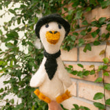 A handcrafted white duck wool felt finger puppet, part of a "Duck Couple on Honeymoon" set, is held by a light-skinned hand against lush greenery and a rustic brick wall. This dapper felt puppet features a textured, bright yellow beak and feet, a small black hat, and a tie. Its charming design highlights the unique, fair-trade craftsmanship, making it ideal for storytelling and imaginative play.
