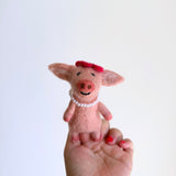 A pink wool felt pig finger puppet, part of a handcrafted piggy couple set, is held upright by a female hand against a plain white background. This charming pig finger puppet is adorned with a red beret and a strand of white pearls around its neck, showcasing its detailed, fair-trade craftsmanship. The hand features red nail polish, complementing the puppet's playful attire.