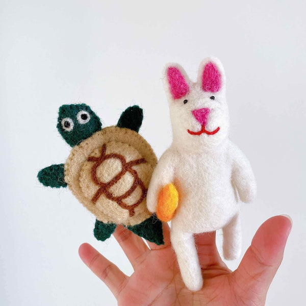 A person's hand holds a set of two handcrafted felt finger puppets, a turtle and a rabbit, against a clean, white background. The dark green felt turtle finger puppet has a tan shell with brown markings. The white felt rabbit finger puppet features pink inner ears, a nose, a stitched red smile, and holds a small orange object. These unique, handmade felt finger puppets are perfect for imaginative storytelling.
