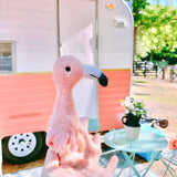A handcrafted felt flamingo finger puppet, part of a set of 5 bird designs, is being held in front of a pink trailer with a white roof parked on grass with trees in the background. The flamingo puppet features a large beak, two eyes, and simple wing patterns. Made from high-quality wool felt, this unique finger puppet showcases intricate detailing and whimsical charm.