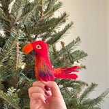 A hand holds a small, colorful felt bird finger puppet against the backdrop of a Christmas tree. The puppet has vibrant plumage with shades of red and orange, giving it a festive appearance. It features detailed feather patterns and a friendly face. The background includes green pine needles adorned with small lights, creating a warm and cozy festive atmosphere.