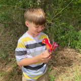 A young boy stands amidst trees and bushes, holding up a small felt finger puppet resembling a bird. The bird has vibrant plumage colors of yellow with blue accents, featuring a large beak and two eyes. This handmade wool felt seagull finger puppet is crafted in Nepal from high-quality materials, showcasing intricate detailing and whimsical charm.