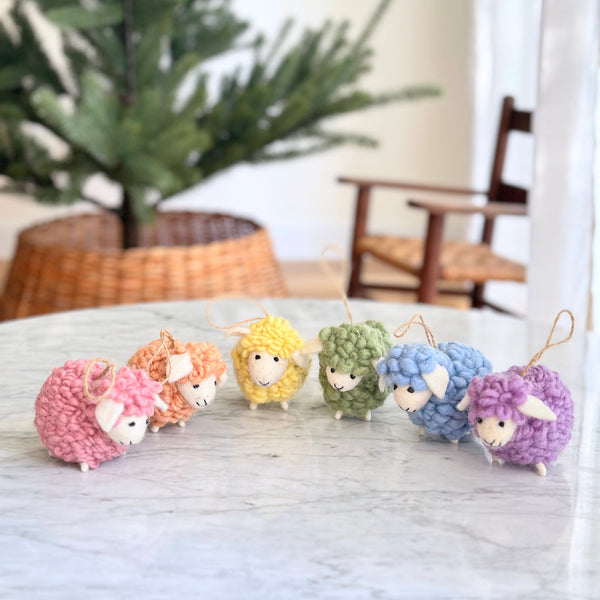 A playful set of 6 pastel-toned felted wool fluffy sheep ornaments is displayed on a marble surface, with a Christmas tree in a basket and a chair in the background. Each mini sheep ornament features a fluffy texture in pink, beige, yellow, green, blue, or purple, and includes a jute string for easy hanging.
