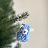 A pastel blue wool felt fluffy sheep Christmas ornament hangs from a green pine tree branch, creating a joyful and festive scene against a soft white background. This charming handmade felt sheep ornament features a fluffy blue body, small white horns, and a simple stitched face, with a jute string attached for hanging.