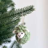 A green felted wool sheep ornament from a pastel tone set hangs from a green pine branch. This handmade felt fluffy sheep ornament features a rounded body with a fluffy texture, white accents on its face and ears. It has small black eyes, a stitched Y-shaped mouth, and a jute string looping over the branch. The unique Christmas ornament evokes a cheerful, festive mood.