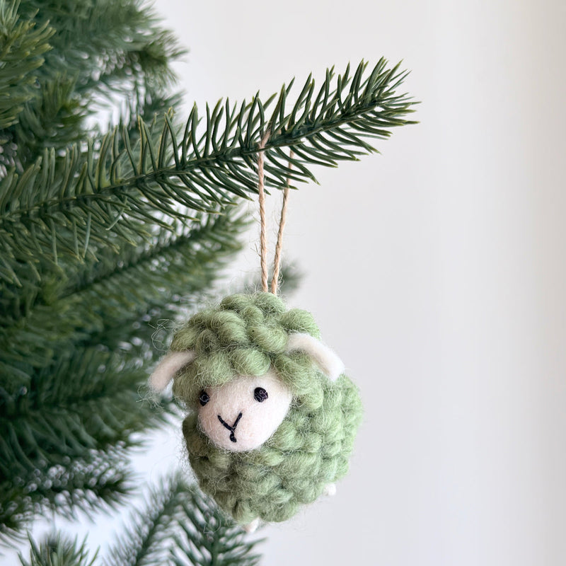 A green felted wool sheep ornament from a pastel tone set hangs from a green pine branch. This handmade felt fluffy sheep ornament features a rounded body with a fluffy texture, white accents on its face and ears. It has small black eyes, a stitched Y-shaped mouth, and a jute string looping over the branch. The unique Christmas ornament evokes a cheerful, festive mood.
