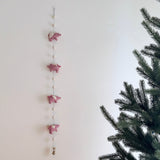 A whimsical felt garland featuring pink flying pigs, suspended against a plain light background, evoking a playful and festive atmosphere. The wool felt garland is adorned with white details, adding a touch of charm to the design. Perfect for decorating nurseries or kids' rooms, this handmade garland exudes a sense of wonder and magic.