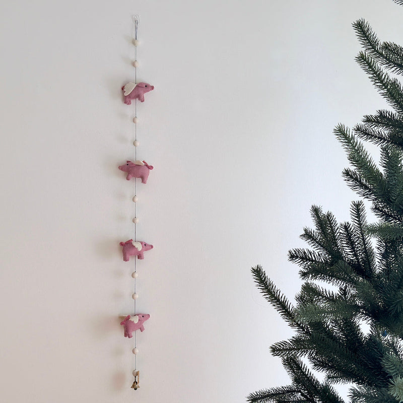 A whimsical felt garland featuring pink flying pigs, suspended against a plain light background, evoking a playful and festive atmosphere. The wool felt garland is adorned with white details, adding a touch of charm to the design. Perfect for decorating nurseries or kids' rooms, this handmade garland exudes a sense of wonder and magic.