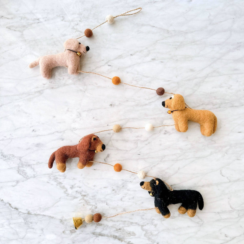 A collection of four dog-shaped felt garlands, each featuring a different color and pattern, are arranged in a straight line on a plain white background. The first dog is cream-colored, the second khaki, the third brown, and the fourth black. Each dog has simple shapes and minimal detailing, with a string or cord connecting some of them.