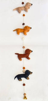 A colorful felt garland featuring four Dachshund puppies, displayed on a white background within a white felt frame. The garland is crafted with cream, khaki, brown, and black felt puppies in various colors, arranged in a line with a felt bone at the center. Made from wool felt, this playful and whimsical garland is perfect for decorating a child's room or as a thematic accessory.