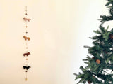 A festive Christmas scene features a string of wool felt garland with four dachshund dogs, each in a different color: cream, khaki, brown, and black. A felt bone charm is attached to the center of the garland, displayed on a white wall.