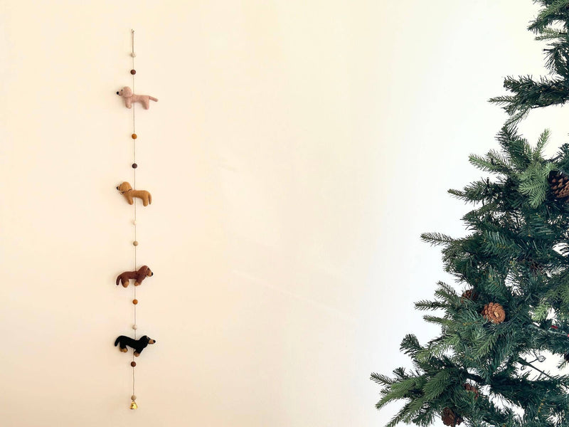 A festive Christmas scene features a string of wool felt garland with four dachshund dogs, each in a different color: cream, khaki, brown, and black. A felt bone charm is attached to the center of the garland, displayed on a white wall.