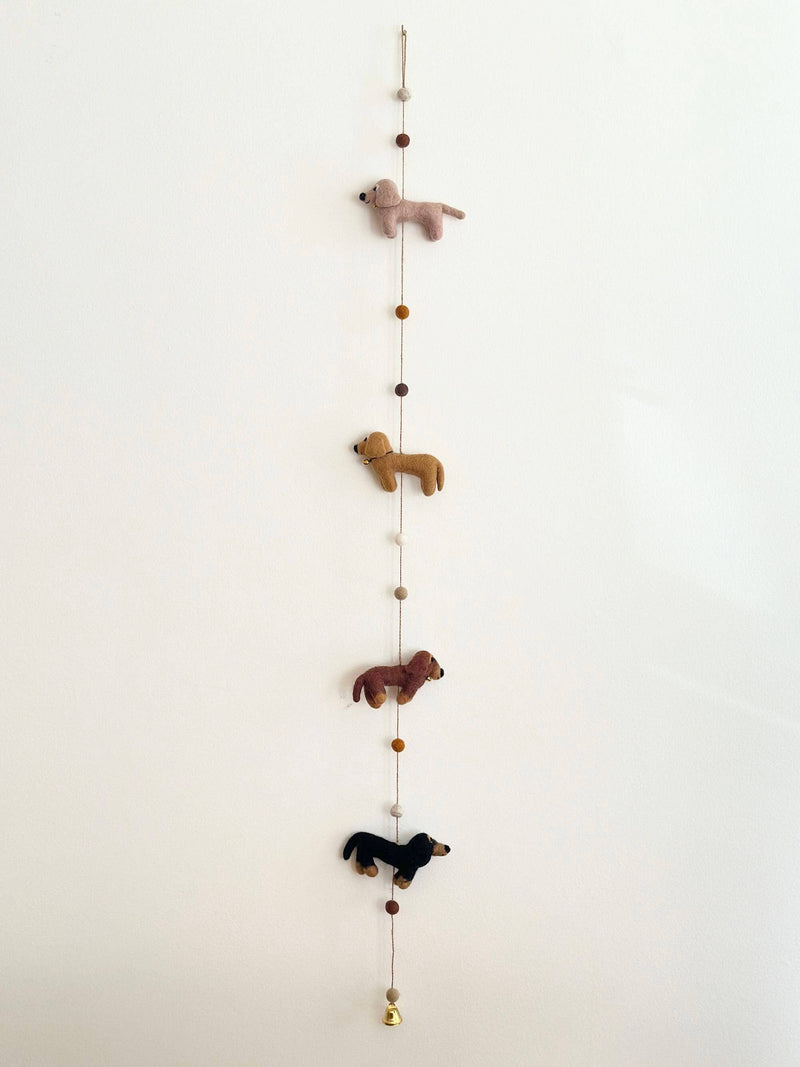 A colorful felt garland featuring four adorable dachshunds in cream, khaki, brown, and black, arranged in a line with the largest at the center. A felt bone is also present on the garland, set against a plain white background.