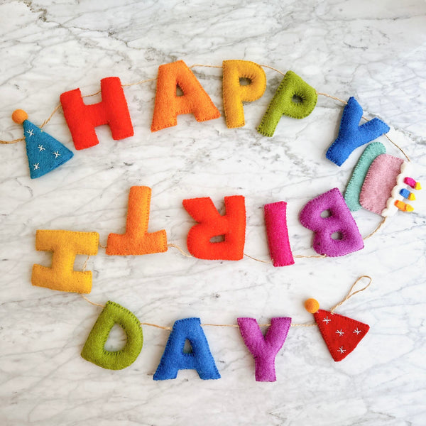 A colorful wool felt 'Happy Birthday' garland rests on a white marble surface. The handcrafted felt garland features vibrant rainbow-toned letters spelling 'HAPPY BIRTHDAY'. Whimsical red and blue party hats, adorned with white snowflakes, flank each end. A charming cake with pink, green, and white layers and colorful candle accents completes the festive design.