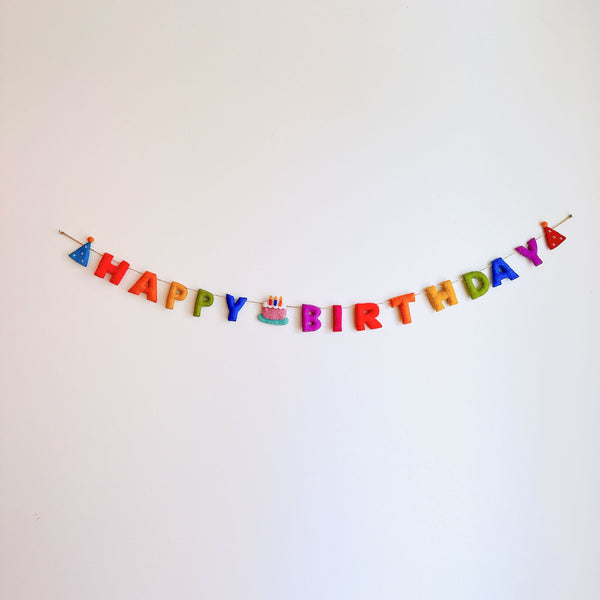 A Happy Birthday wool felt garland is displayed against a stark white backdrop, evoking a cheerful and celebratory atmosphere. This handcrafted felt garland spells "HAPPY BIRTHDAY" in vibrant rainbow letters: red, orange, yellow, green, blue, and purple. The string features whimsical party hats on each end and a delightful cake with a candle in the middle, enhancing its festive appeal.