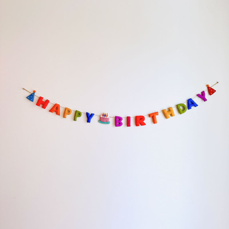 A Happy Birthday wool felt garland is displayed against a stark white backdrop, evoking a cheerful and celebratory atmosphere. This handcrafted felt garland spells "HAPPY BIRTHDAY" in vibrant rainbow letters: red, orange, yellow, green, blue, and purple. The string features whimsical party hats on each end and a delightful cake with a candle in the middle, enhancing its festive appeal.