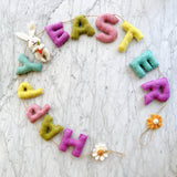 A circular felt garland with pastel-colored letters spelling out 'Happy Easter' is displayed on a marble countertop, surrounded by decorative elements like daisies and bunnies holding carrots. The garland measures 48 inches long and features whimsical pastel hues, making it perfect for Easter celebrations. Crafted from durable jute thread and natural New Zealand sheep wool felt, this unique garland adds a playful touch to any room.