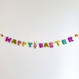 A wool felt garland hangs on a white wall, spelling out 'Happy Easter' in pastel colors. The garland features decorative elements like daisies, sunflowers, and bunnies with carrots, showcasing handcrafted details from natural sheep wool materials.