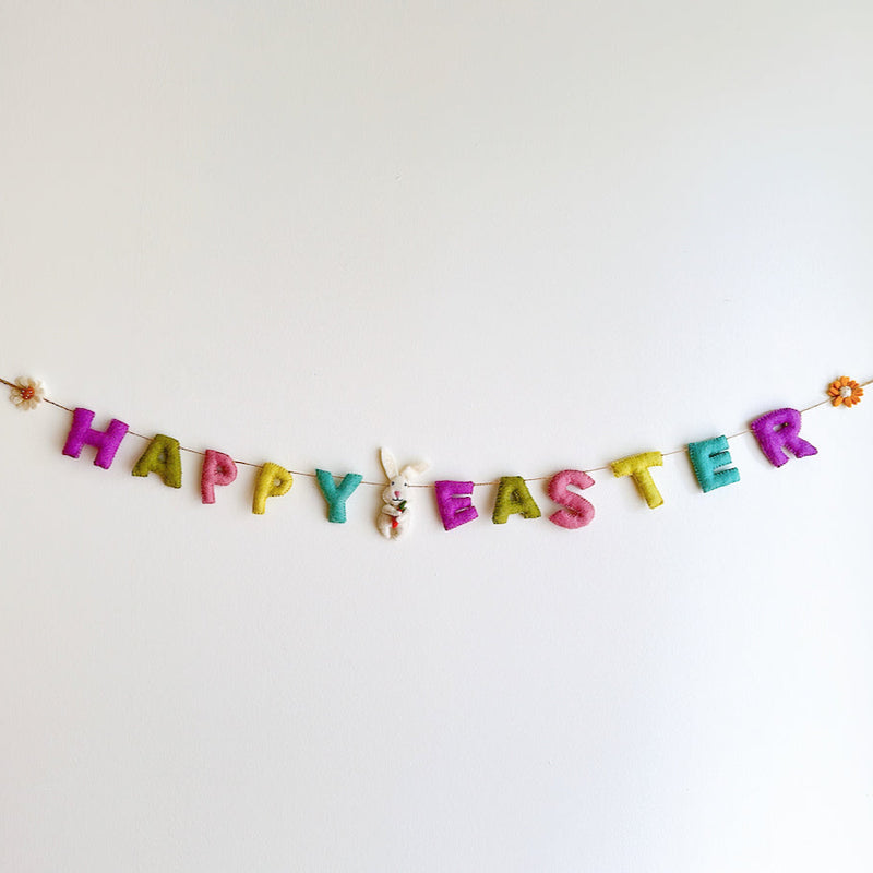 A wool felt garland hangs on a white wall, spelling out 'Happy Easter' in pastel colors. The garland features decorative elements like daisies, sunflowers, and bunnies with carrots, showcasing handcrafted details from natural sheep wool materials.