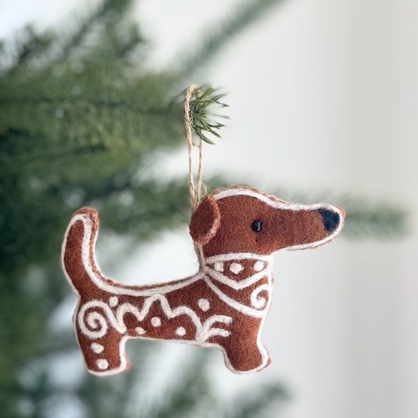 A needle felted Gingerbread Dachshund Dog ornament hangs against a blurred Christmas tree, evoking a festive and joyful mood. This brown felt ornament, resembling a gingerbread cookie, features white icing-like patterns, small round ears, a short tail, and intricate icing swirls and dots. A jute string allows it to hang gracefully.