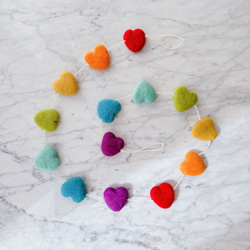 A colorful wool felt star garland is cheerfully displayed on a white marble-patterned surface. The garland features multiple soft, textured stars in a pastel rainbow of red, orange, yellow, green, blue, and purple. Each star is connected by a thin white cotton thread.