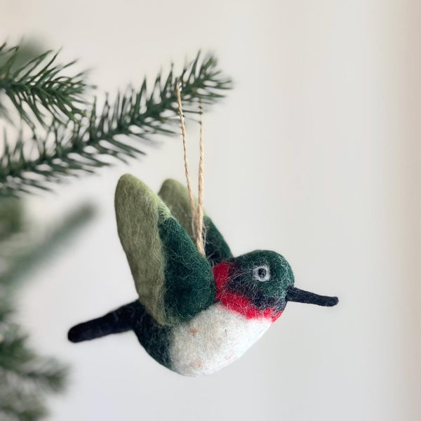 A needle felted wool hummingbird Christmas ornament hangs gracefully from a green pine branch, suggesting a festive tree setting. This charming 3D felt hummingbird features a dark green head, a vibrant red throat patch, a white chest, and shaded green wings. It has a long black beak and a tiny bead eye, complete with a jute string for easy display.
