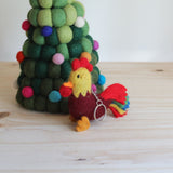 A handcrafted rooster wool felt keychain rests on a wooden surface beside a decorative felted green Christmas tree. The rooster keychain features a burgundy body, red and yellow accents, an orange beak, and vibrant red, blue, green, and yellow tail feathers, complete with a metal clasp. The festive setting includes a green felt Christmas tree adorned with small colorful felt sphere ornaments.
