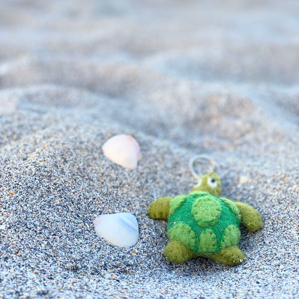 A handcrafted sea turtle wool felt keychain rests on a sandy surface with scattered seashells, evoking a calm, coastal charm. This unique felt keychain features a darker green head and limbs, a lighter green shell with darker green circular details, and a metal keychain.