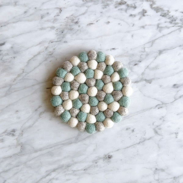 A set of four felt light blue round coasters rests on a white marble surface with subtle gray veining. Each handcrafted felt coaster is made from small, hand-stitched balls of 100% organic lamb wool in varying shades of light blue. Ethically handmade by Nepalese female artisans, these unique wool felt coasters offer a calm, minimalist aesthetic.