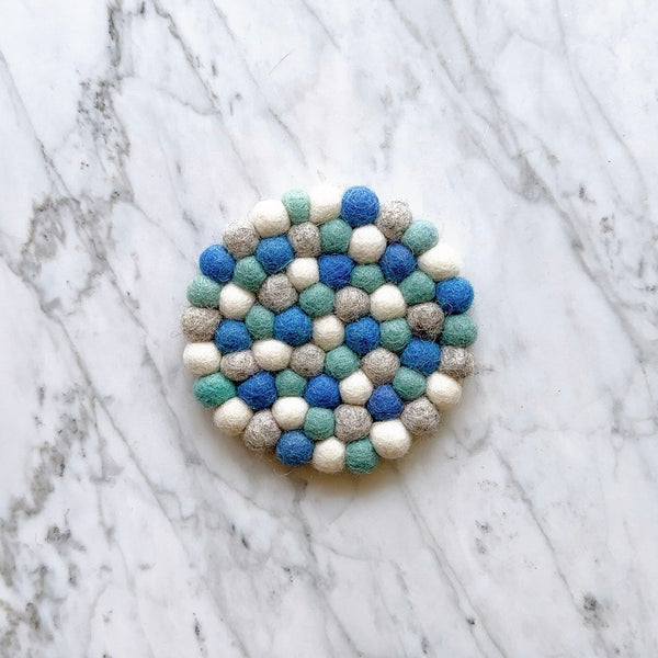 A single organic wool felt coaster in light gray and ocean blue lies flat on a white marble-like surface. This round, handcrafted felt coaster features small, tightly packed, hand-stitched felt balls, highlighting its unique texture and ethically made quality.