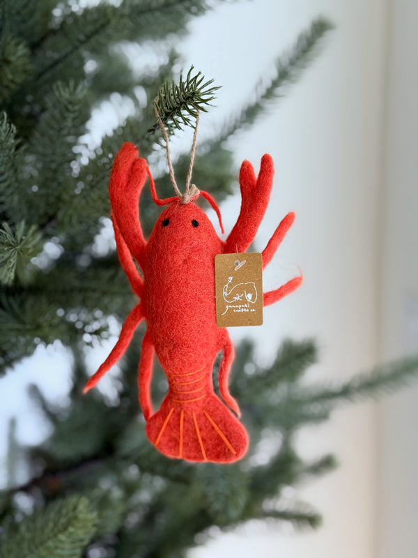 A vibrant red wool felt lobster ornament hangs from a lush green Christmas tree. The handcrafted felt lobster features detailed claws, antennae, and a jute string attached for easy hanging, adding a cheerful, nautical touch to the holiday decor.