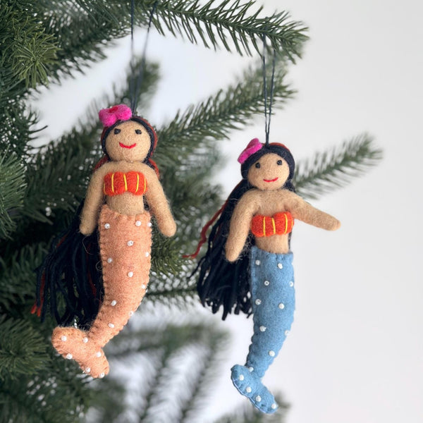 Two handmade wool felt mermaid Christmas ornaments with black hair hang on a lush green Christmas tree, set against a clean white background. Each felt mermaid ornament features a tan skin tone, an orange top, and black hair with a small pink flower. One mermaid has a peach-colored tail, and the other a blue tail, both adorned with white dots and ready to hang with a black string.