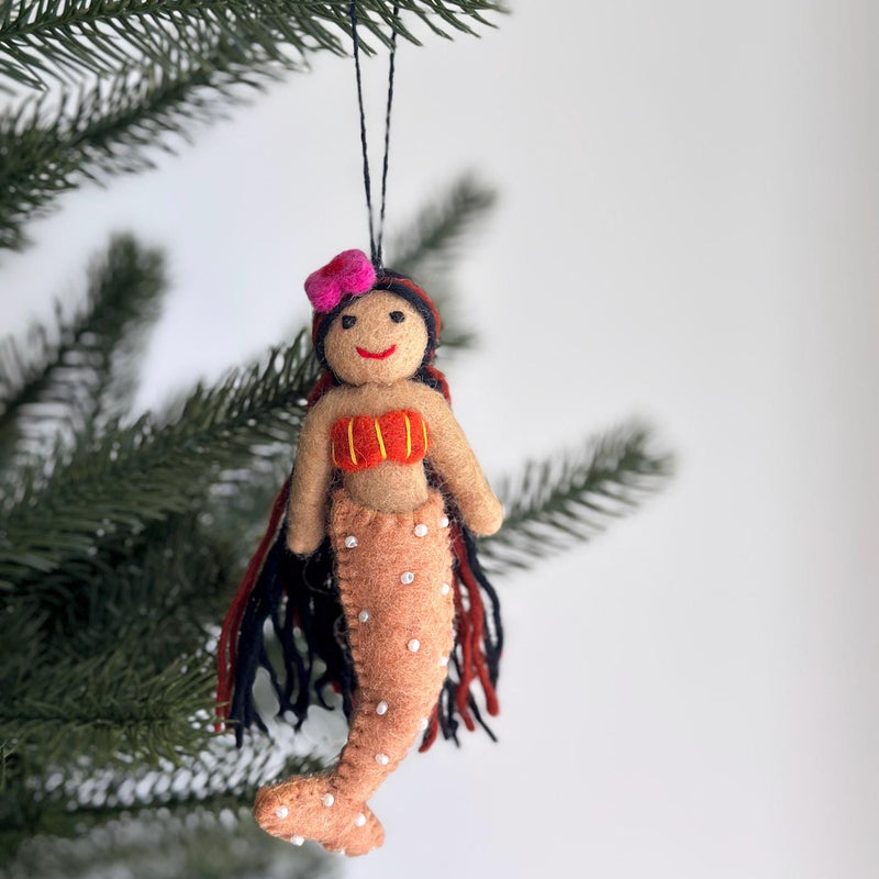 A felt mermaid Christmas ornament with black hair is seen in a close-up, hanging on a Christmas tree branch. The unique wool felt mermaid features tan skin, long red and black hair, a pink flower on its head, a bright red bra, and an orange tail adorned with small white beads. This needle-felted, 3D designed ornament has a black string for easy hanging.