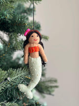 A handmade wool felt mermaid ornament with black hair hangs from a vibrant green Christmas tree branch. This detailed Christmas ornament features a black cotton string for hanging, a pink flower in her hair, a bright orange top, and a pale green tail embellished with small white beads, highlighting its unique, festive charm.