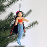 A wool felt mermaid Christmas ornament with black hair hangs from a blurred Christmas tree. This unique handmade felt ornament features a light tan body with a long blue tail adorned with white dots. She has black hair with red accents, a pink hat, a smiling red mouth, and an orange top with yellow stitching.
