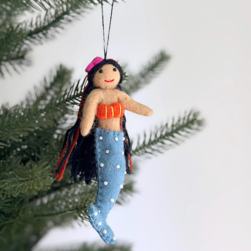 A wool felt mermaid Christmas ornament with black hair hangs from a blurred Christmas tree. This unique handmade felt ornament features a light tan body with a long blue tail adorned with white dots. She has black hair with red accents, a pink hat, a smiling red mouth, and an orange top with yellow stitching.