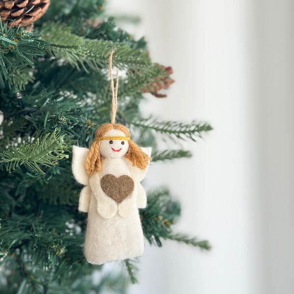 A handmade felt mini angel Christmas ornament hangs from a lush green Christmas tree branch. The white wool felt angel features light brown hair, a gold head band, and a brown heart sewn onto its body. A sweet red stitched smile adds charm, and it is suspended by a natural jute string.