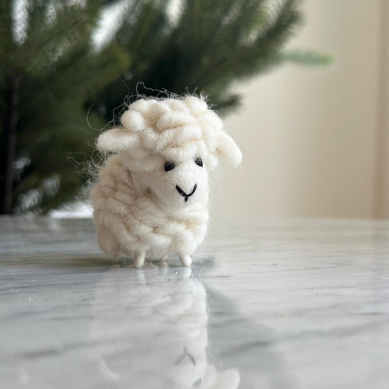A cute felt mini sheep ornament, ideal as a table decoration, is displayed on a glossy marble surface before a blurred green tree. This charming handmade ornament, crafted from fluffy off-white wool, features a rounded body, small black eyes, and a stitched smiling mouth. It wears a colorful scarf and has delicate thin legs, with a hemp string loop for hanging.