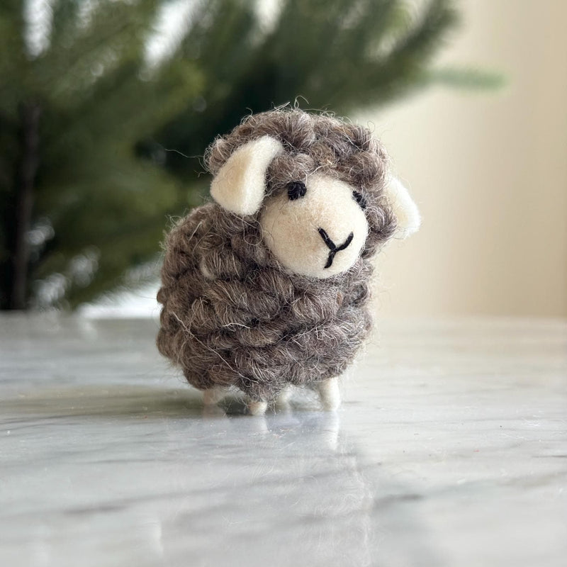 A charming felt mini sheep ornament sits on a white marble surface, against a softly blurred evergreen tree and a light beige wall. This handmade wool felt ornament features a textured body of mixed brown and white, with small white felt ears and legs. Its smooth white felt face has simple black eyes and a stitched 'Y' mouth, complete with a hemp string loop for hanging.