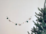 A wool felt mountain and cloud garland is displayed against a white wall, with a large pine tree partially visible on the right. The handcrafted garland features alternating 3D wool felt shapes of dark green pine trees, grey and white mountains, and fluffy white clouds. Small grey and dark green felt balls separate these elements along a white cord.