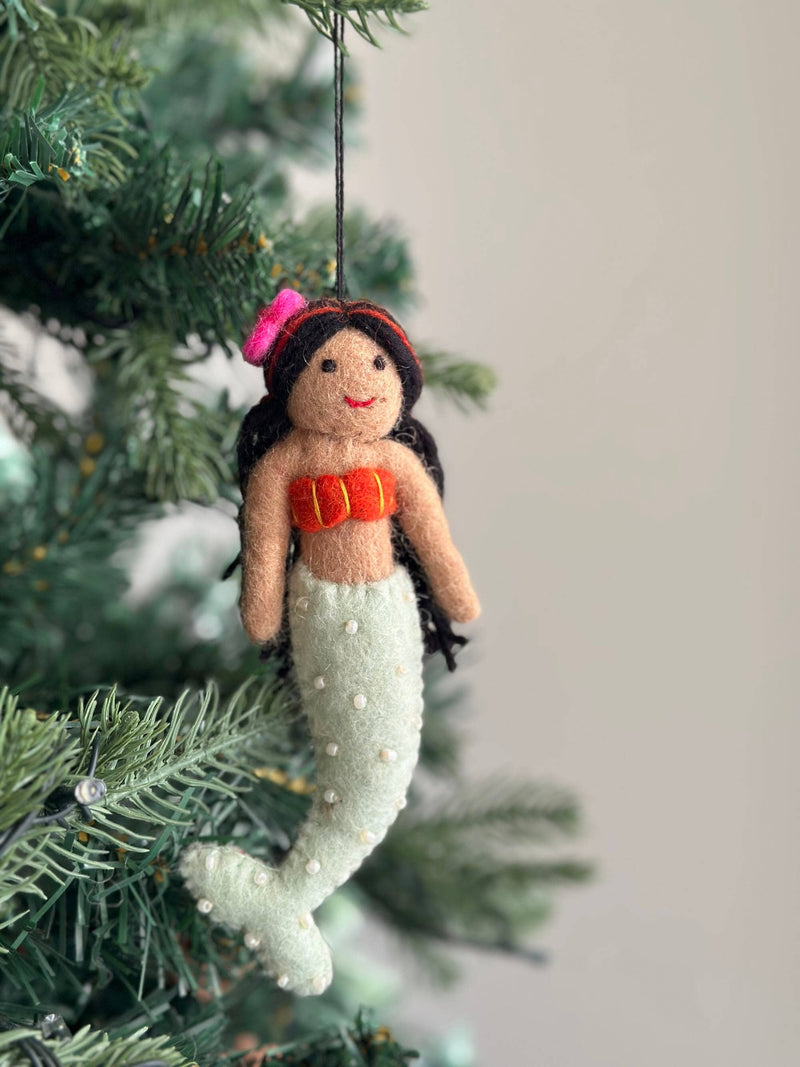 A wool felt mermaid ornament hangs on a Christmas tree branch, creating a playful, festive atmosphere. This unique handmade ornament features a tan upper body, a bright red top with yellow stripes, and a light green tail adorned with small white beads. The mermaid has black hair with a pink flower and bendable limbs for posing. A black string is attached for hanging.