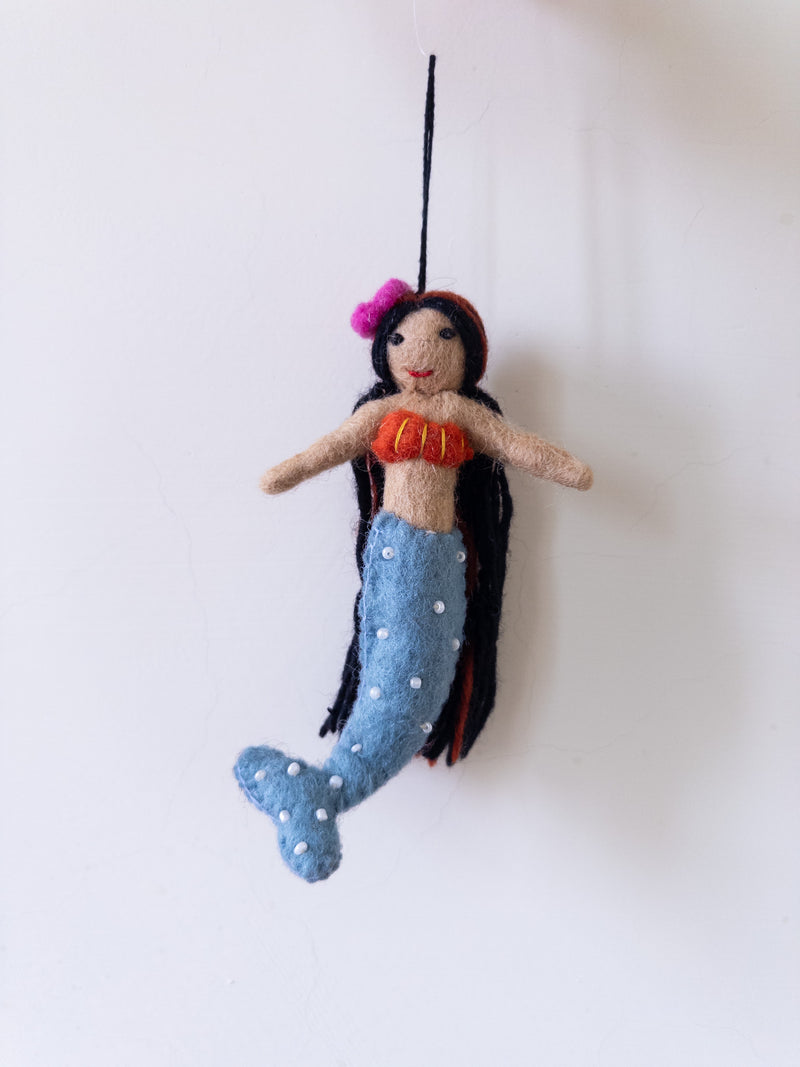 A handmade wool felt mermaid ornament is suspended against a plain white backdrop, creating a whimsical scene. The felt mermaid features a tan upper body with bendable outstretched arms and an orange top with yellow stripes. Her long black hair is accented with a pink flower, complementing her bendable blue tail adorned with white dots. A black cotton string is attached for easy hanging.